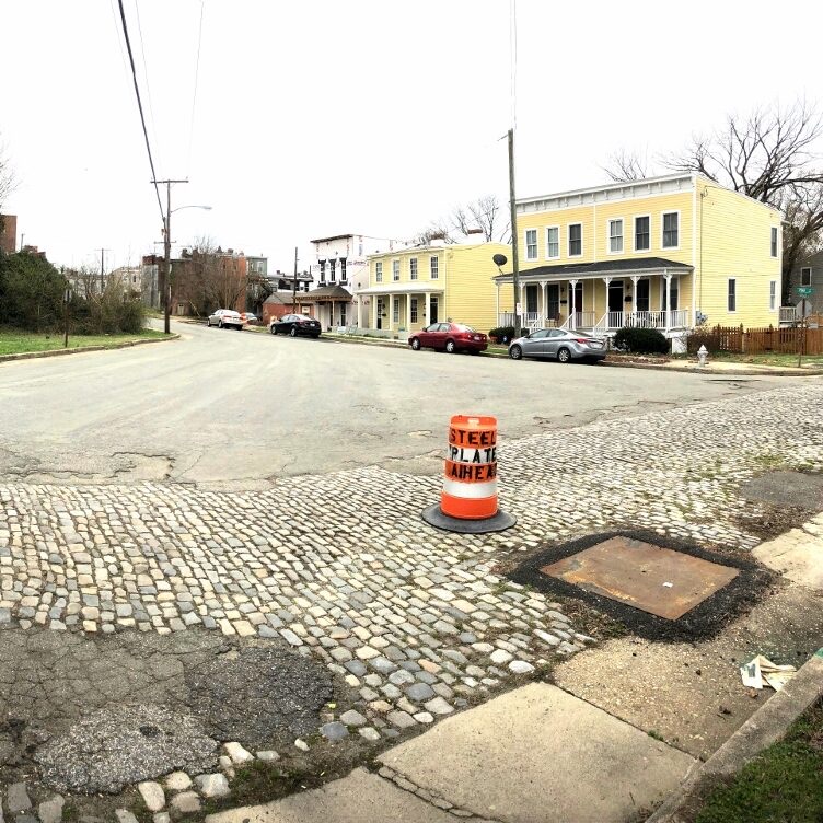 Cobblestones and asphalt patches at the corner of Pink Street and Carrington, with a steel-plate warning barrel